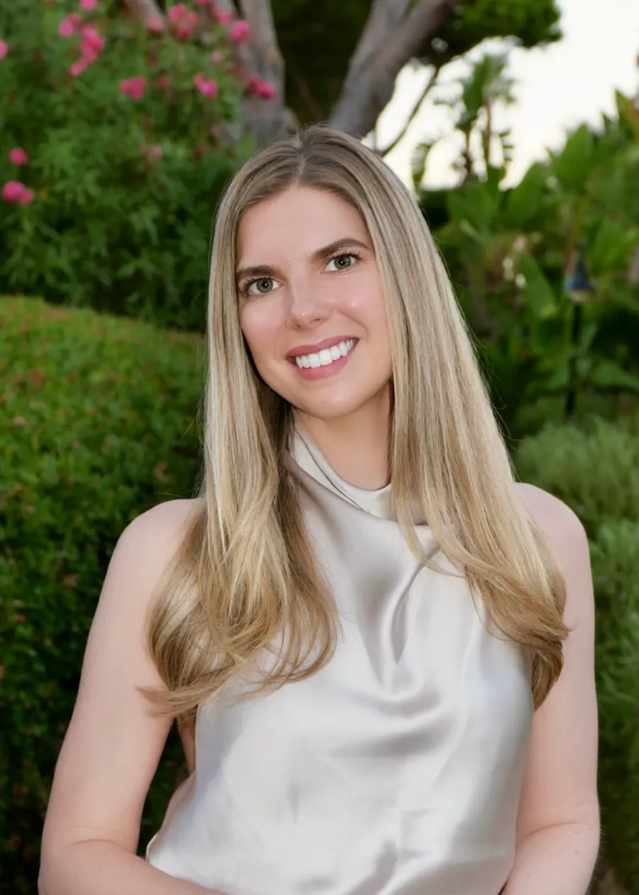 Photograph of Samantha Jane Lever smiling, her long blonde hair over a pale satin top, in a garden of green hedges and pink flowers
