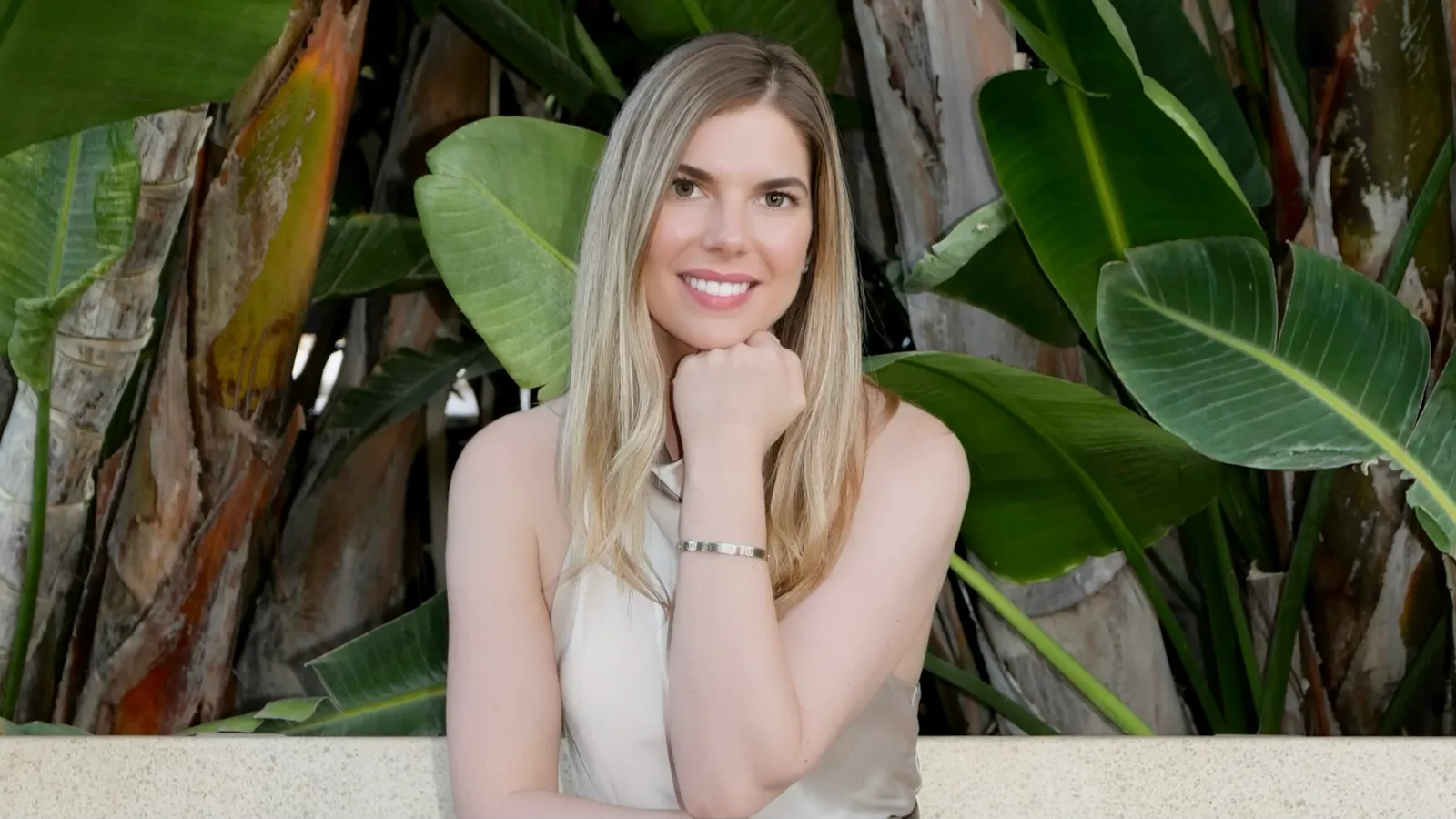 Photograph of Samantha Jane Lever smiling, chin resting on her hand, in front of large green banana leaves