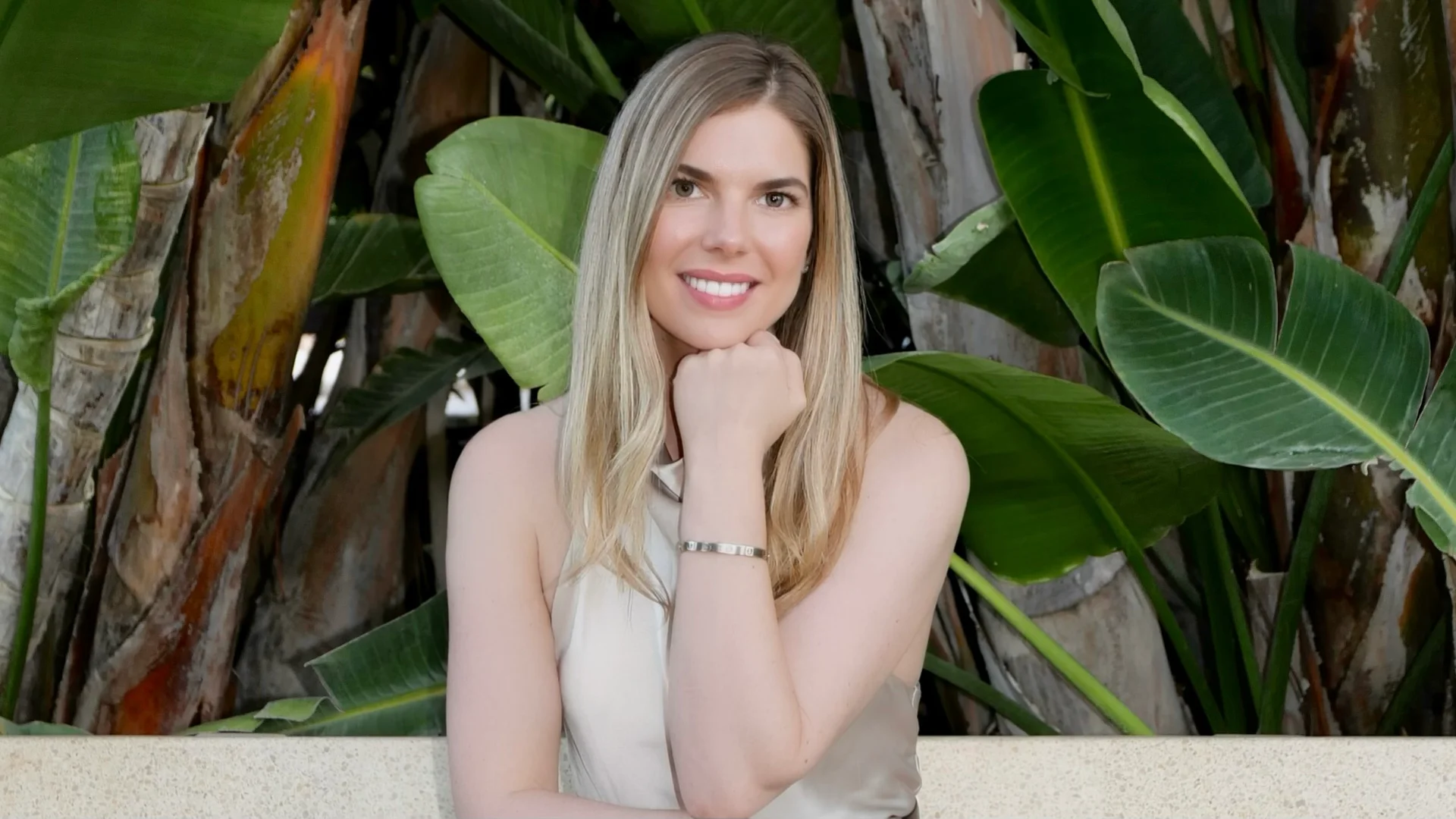 Photograph of Samantha Jane Lever smiling, chin resting on her hand, in front of large green banana leaves